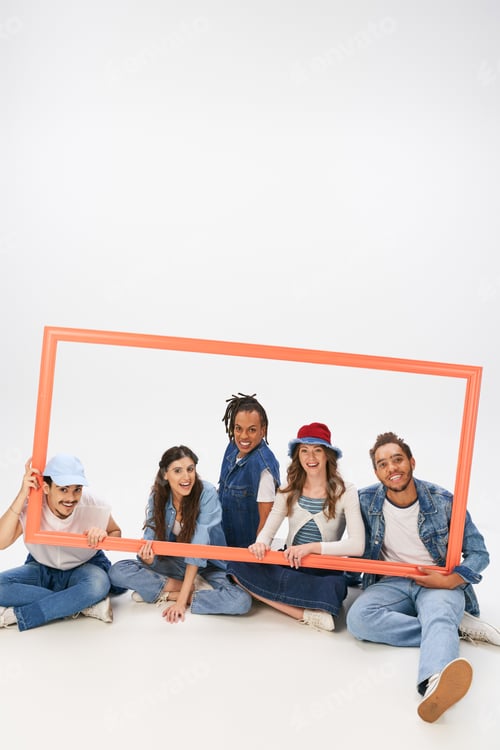 Preview: joyful and trendy multiethnic friends sitting and posing with picture frame on grey backdrop
