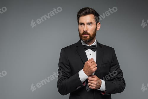 Preview: bearded man in black tuxedo with bow tie adjusting sleeve on white shirt isolated on grey