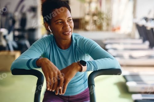 Preview: Black athletic woman taking a break after exercising on stationary bike in gym.