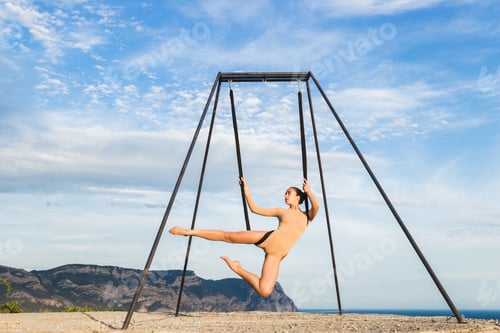Preview: Woman practicing fly-dance yoga poses in a hammock on the coast with views of the mountains