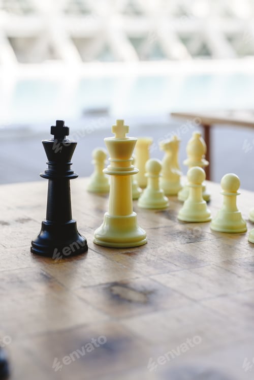 Preview: Black and white chess kings faced between defocused pieces on wooden board.