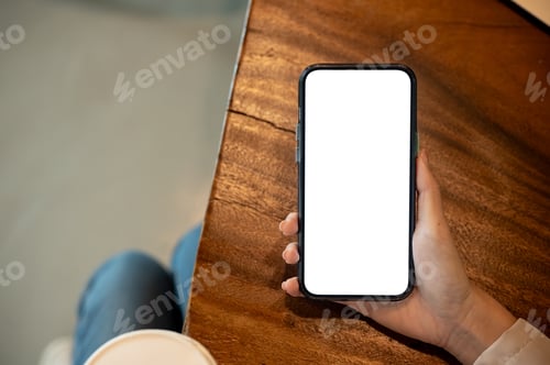 Preview: A woman hand holding blank screen smartphone aside coffee on wooden table while sitting cross legged
