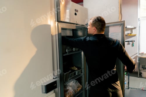 Preview: chef stands in kitchen in a black uniform and looks through what is on the list in the refrigerator