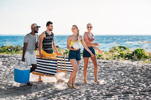 Preview: happy young multiethnic friends with beach chairs, cooler and ball walking on sandy beach