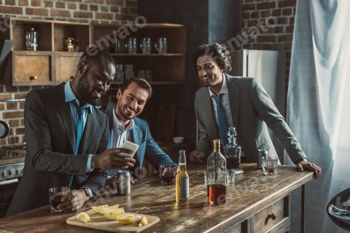 Preview: smiling male friends in suits using smartphone and drinking alcohol beverages while partying at home