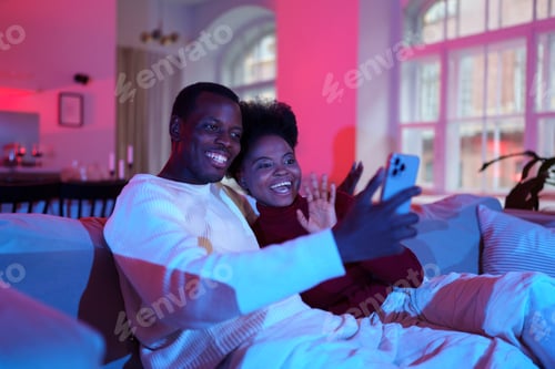 Preview: Cheerful african couple talk by video call on smartphone with friends while relaxing on sofa at home