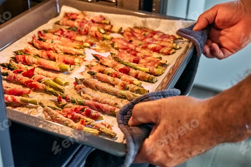 Preview: Hands of man taking baking tray with wrapped asparagus in ham from the oven.