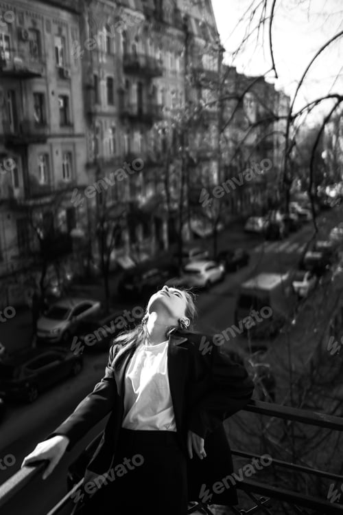 Preview: Black and white photo of stylish woman posing on balcony in city. Spring autumn fashion concept.