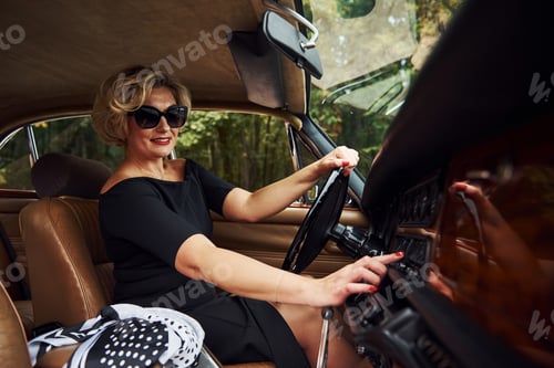 Preview: Blonde woman in sunglasses and in black dress sits in old vintage classic car