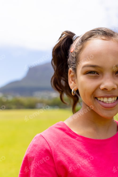Preview: Biracial girl with a bright smile, wearing a pink shirt, stands outdoors with copy space