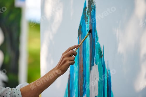 Preview: Artist hand holds paint brush and draws abstract surreal image on white canvas at outdoor festival