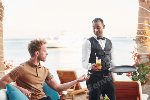 Preview: Serviced by waiter. Young man is outdoors at sunny daytime. Concept of vacation