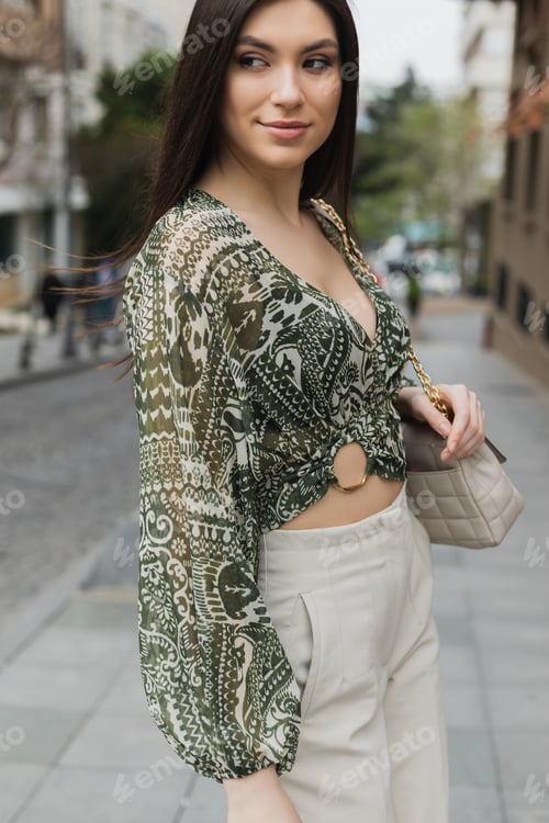 Preview: stylish woman with brunette long hair in trendy outfit with beige pants, cropped blouse and handbag