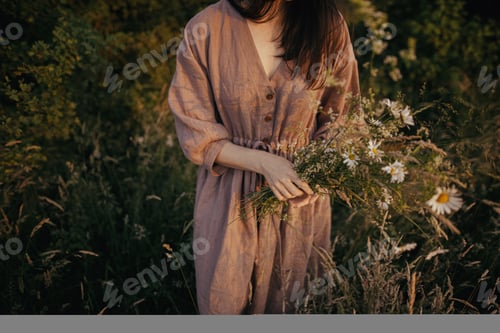 Preview: Beautiful woman in linen dress gathering wildflowers in summer meadow in sunny evening. Slow life