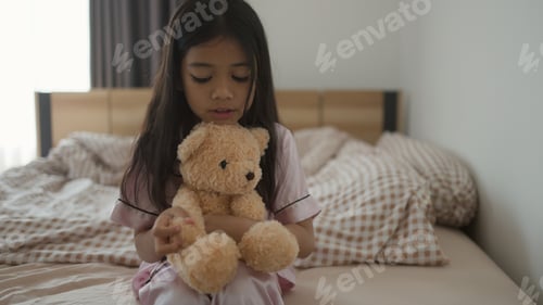 Preview: Sweet Asian little girl is hugging a teddy bear and smiling while sitting on her bed at home