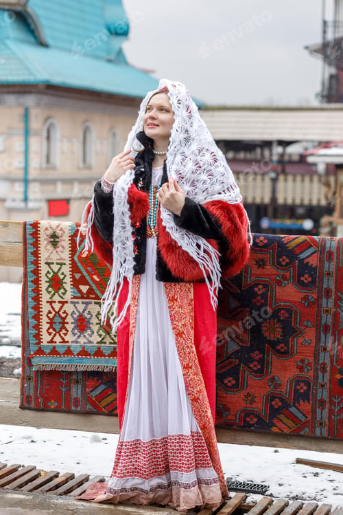 Preview: A Russian girl in red traditional Russian clothes and white shawl near wooden house in spring