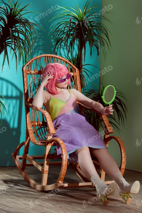 Preview: fashionable young female model in pink wig sitting in rocking chair and looking at mirror