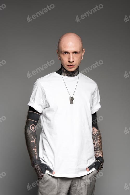 Preview: handsome bald tattooed man in white t-shirt standing with hands in pockets and looking at camera