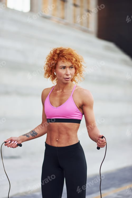 Preview: Young european redhead woman in sportive clothes jumping with rope outdoors