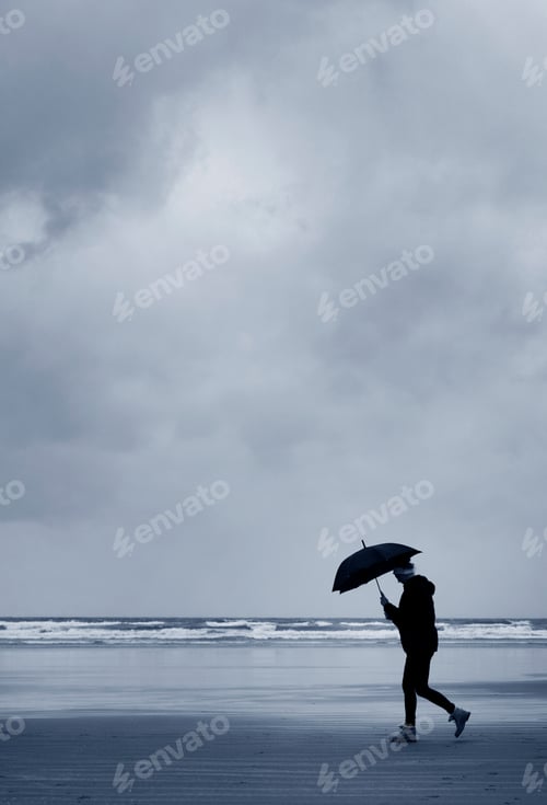 Preview: Side view of man carrying umbrella walking along sandy beach by ocean under stormy sky.