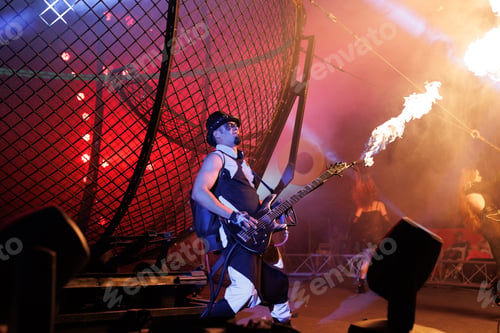 Preview: Male showman in the circus arena in the light of multicolored spotlights with a guitar and fire.