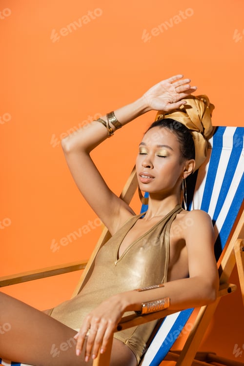 Preview: Fashionable african american model in swimsuit and headband sitting on deck chair on orange