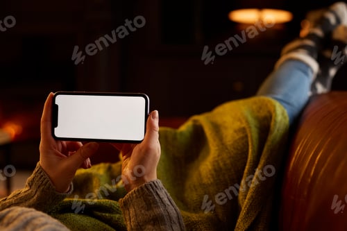 Preview: Close Up Of Woman At Home Lying In Lounge With Cosy Fire Streaming Onto Blank Screen Mobile Phone