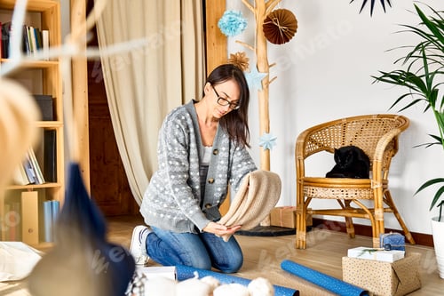 Preview: Woman wrapping woolen sweater in craft recycled paper in the room with alternative christmas tree