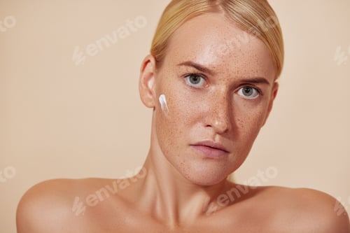Preview: Beautiful blond woman with cream on her face looking straight of a camera against pastel backdrop