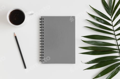 Preview: Top view of a grey notebook mockup with workspace accessories and a palm leaf on a white table.