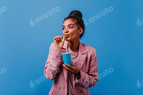 Preview: Dreamy young woman in pink jacket drinking cold tea. Pretty african girl enjoying soda on blue back