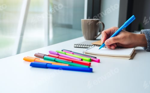 Preview: Closeup image of a woman writing on a blank notebook with colored pens and coffee cup on the table