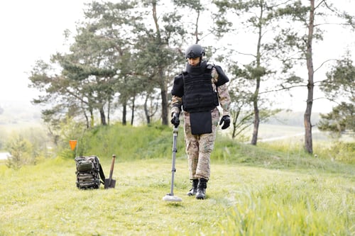 Preview: A man in a military uniform and bulletproof vest works in the forest with a metal detector