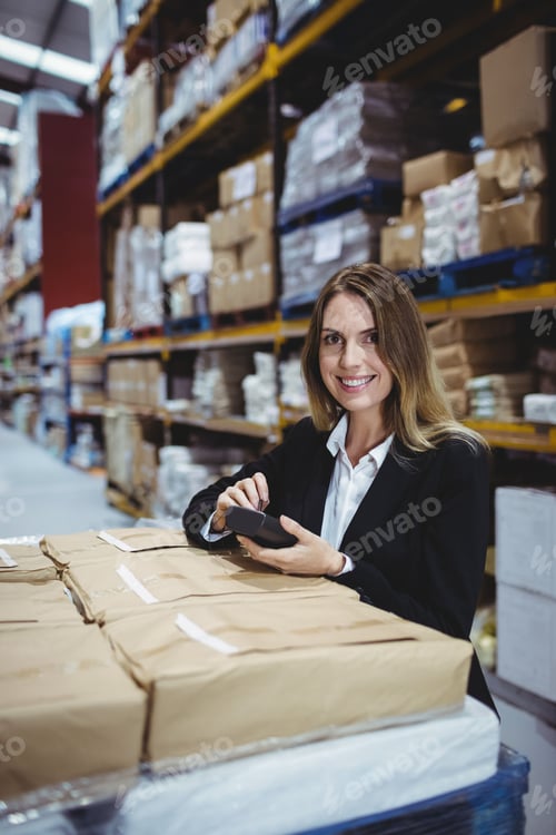 Preview: Woman in business attire standing at large warehouse scanning brown packages with handheld scanner