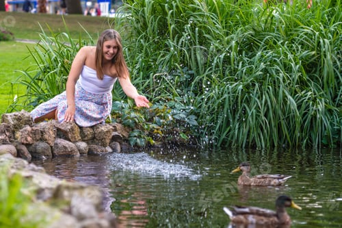 Preview: a young woman sits on rocks by a pond and splashes water on ducks