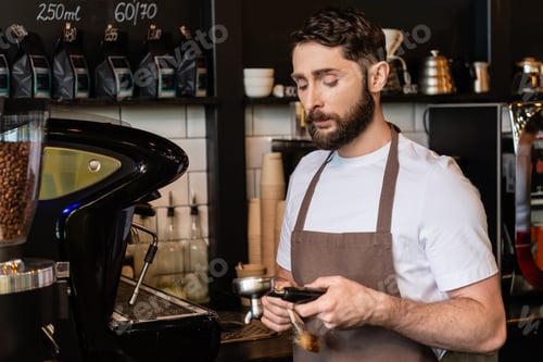 Preview: bearded barista in apron holding brush and coffee machine holder while working in coffee shop
