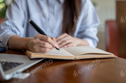 Preview: Close up of blue shirt woman writing in notebook with a fountain pen sitting at cafe's wooden table