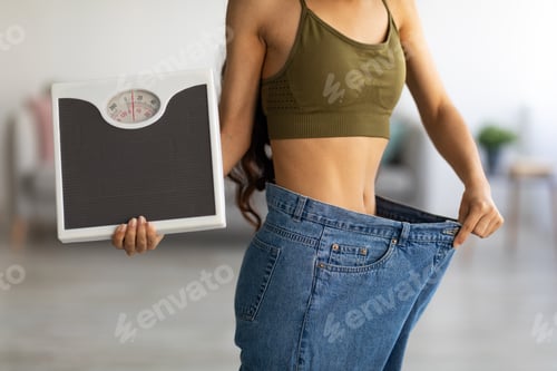 Preview: Closeup of Indian woman in oversized jeans holding scales, showing results of weight loss program or