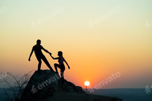 Preview: Man and woman hikers helping each other to climb a big stone at sunset in mountains.