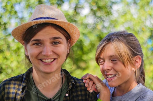 Preview: close-up smiling young brunette caucasian woman in hat with preteen girl. Summer blurred green