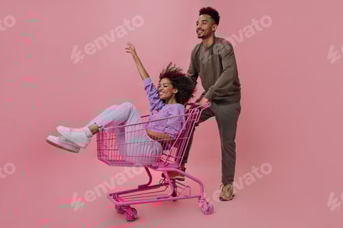 Preview: Man in brown outfit riding girlfriend in shopping cart on isolated. Portrait of guy and teen curly