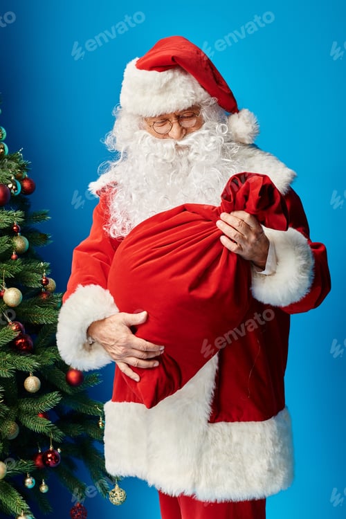 Preview: Santa Claus with beard and eyeglasses looking at red sack bag with Christmas presents on blue