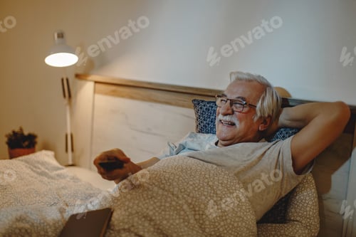 Preview: Happy mature man lying down in bed and watching TV at night.