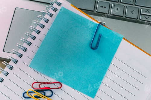 Preview: Top view closeup of a blue sticky note inside of a notebook, secured with a paper clip and a laptop
