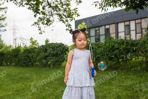 Preview: asian girl in dress holding bubble stick and looking at soap bubble outside