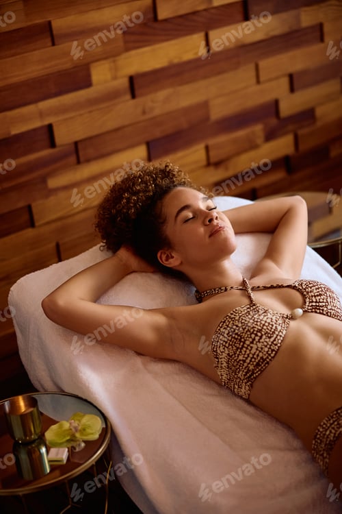 Preview: Young woman with eyes closed relaxing on deck chair at wellness center.