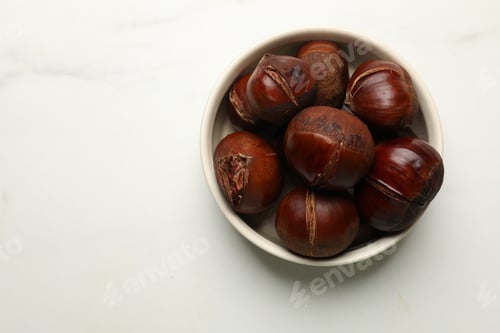 Preview: Roasted edible sweet chestnuts in bowl on white marble table, top view. Space for text