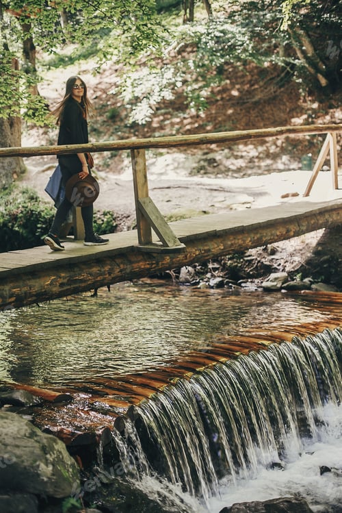Preview: stylish traveler girl with hat walking at waterfall river in sunny forest