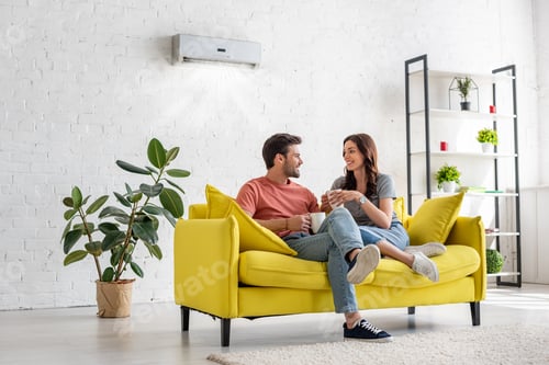Preview: happy young couple talking and holding cups while sitting on yellow sofa under air conditioner at