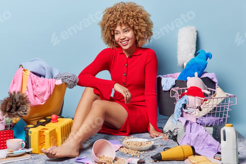 Preview: Cheerful curly lady in elegant red dress poses on floor with different items around pose in messy